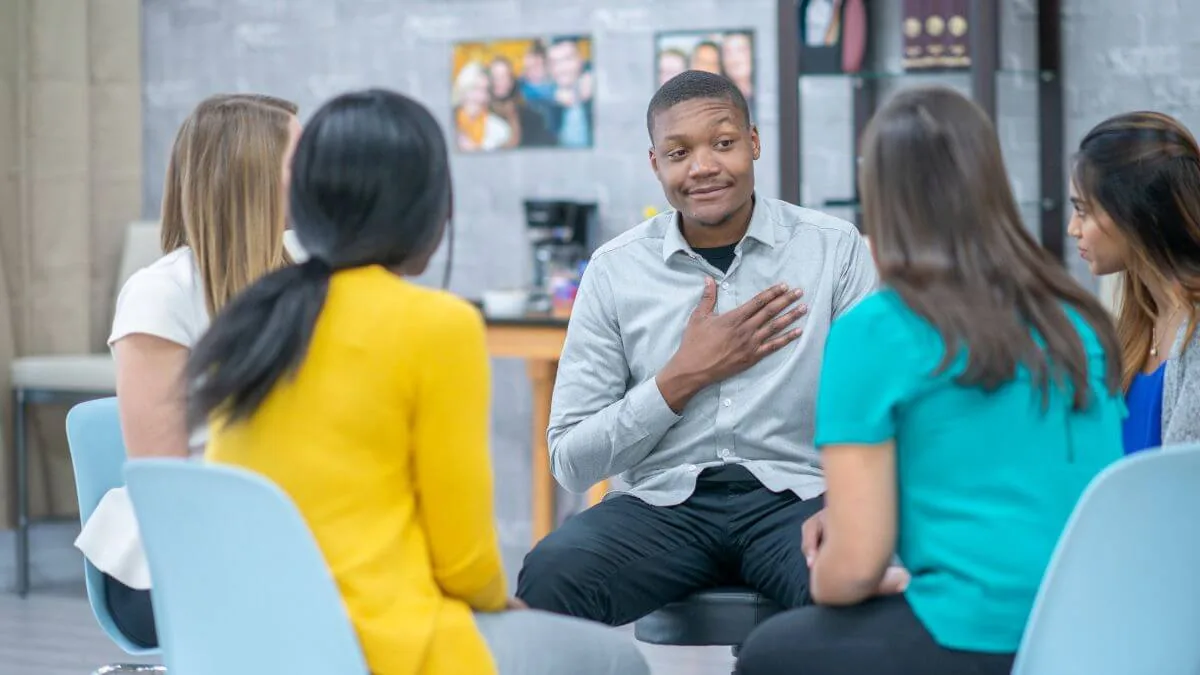 Five people sit in a circle; a man in a gray shirt speaks with his hand on his chest while others listen.