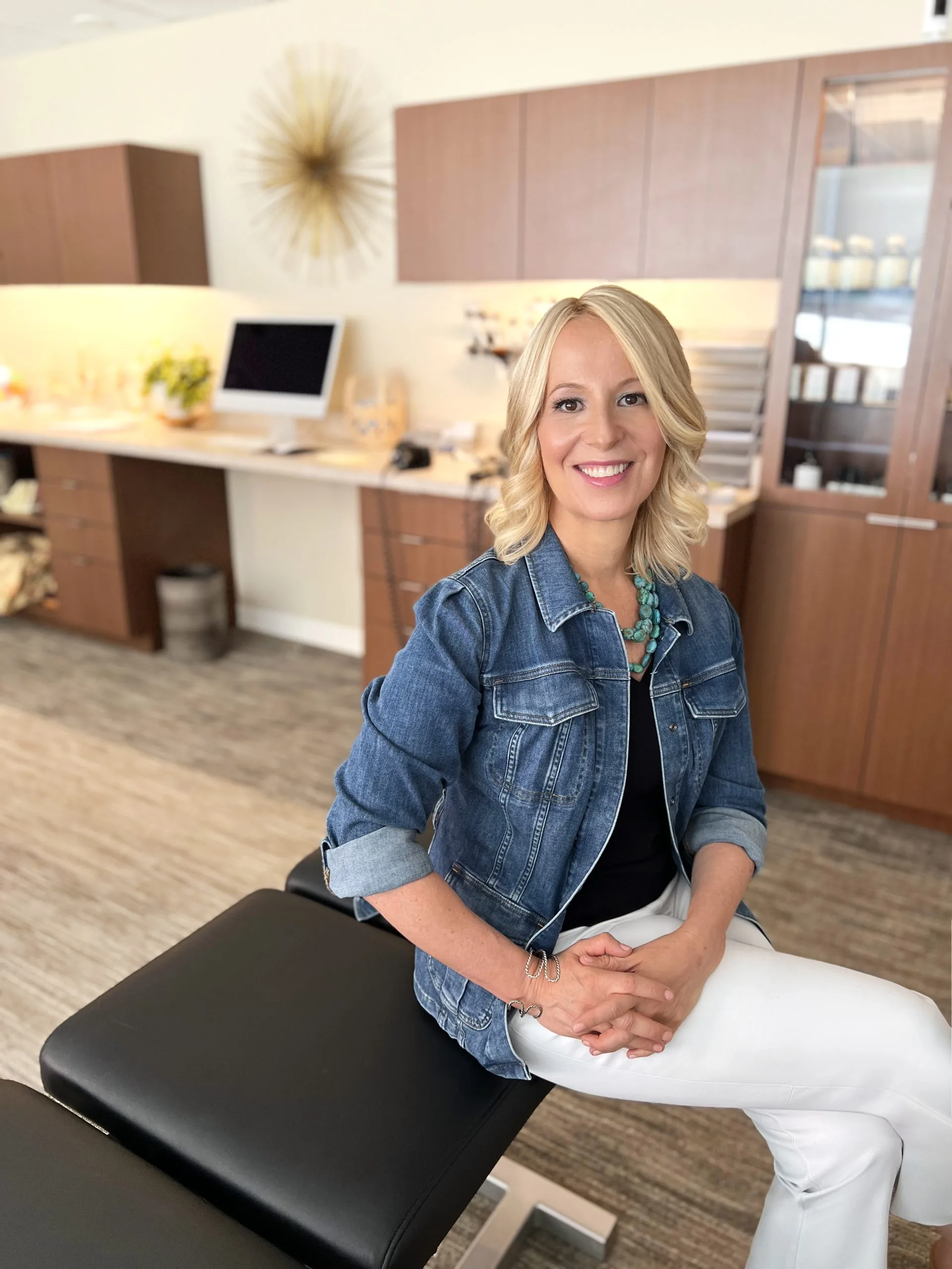 Smiling woman seated on a chiropractic adjustment table in a clinic reception area, wearing a denim jacket and turquoise necklace.