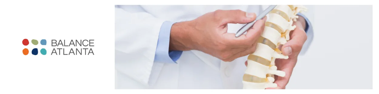 Person in a white coat pointing at a model spine with a pen, Balance Atlanta logo at left.