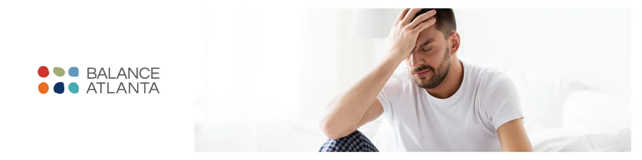 Man sitting on a bed with his hand on his forehead, appearing tired or in discomfort, with the Balance Atlanta logo at left.