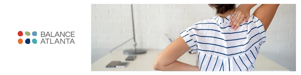 Person seen from behind holding their upper back and neck while seated at a desk, wearing a striped shirt with the Balance Atlanta logo at left.