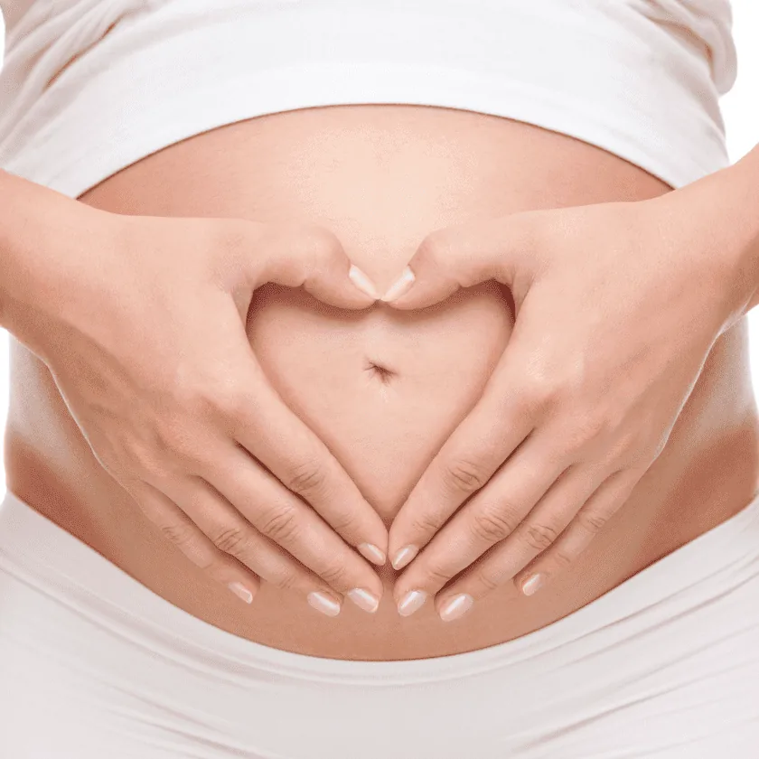 Hands forming a heart shape over a pregnant belly, woman wearing white clothing visible