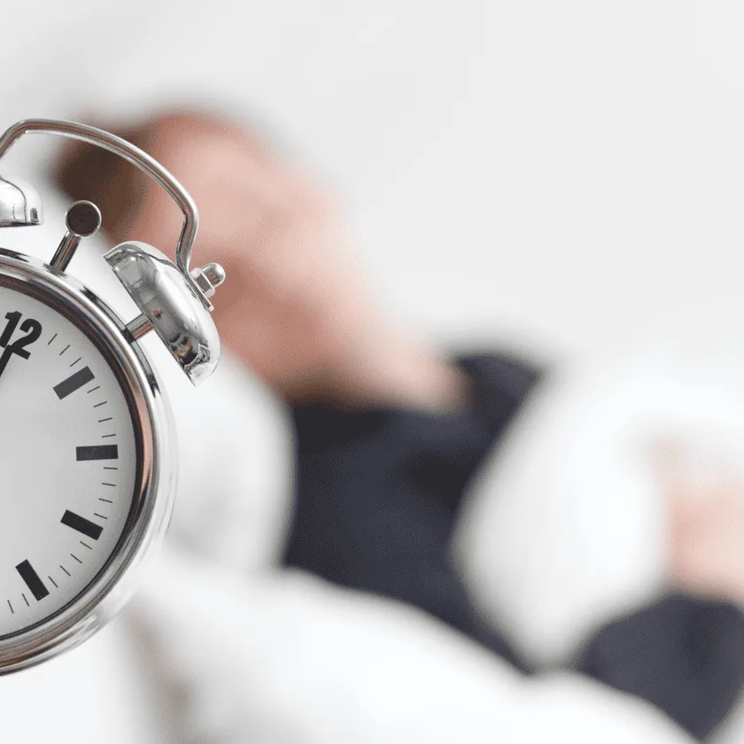 Silver analog alarm clock in the foreground with a blurred person lying in bed in the background