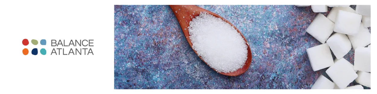 Wooden spoon of granulated sugar and scattered sugar cubes on a blue textured surface, Balance Atlanta logo at left.