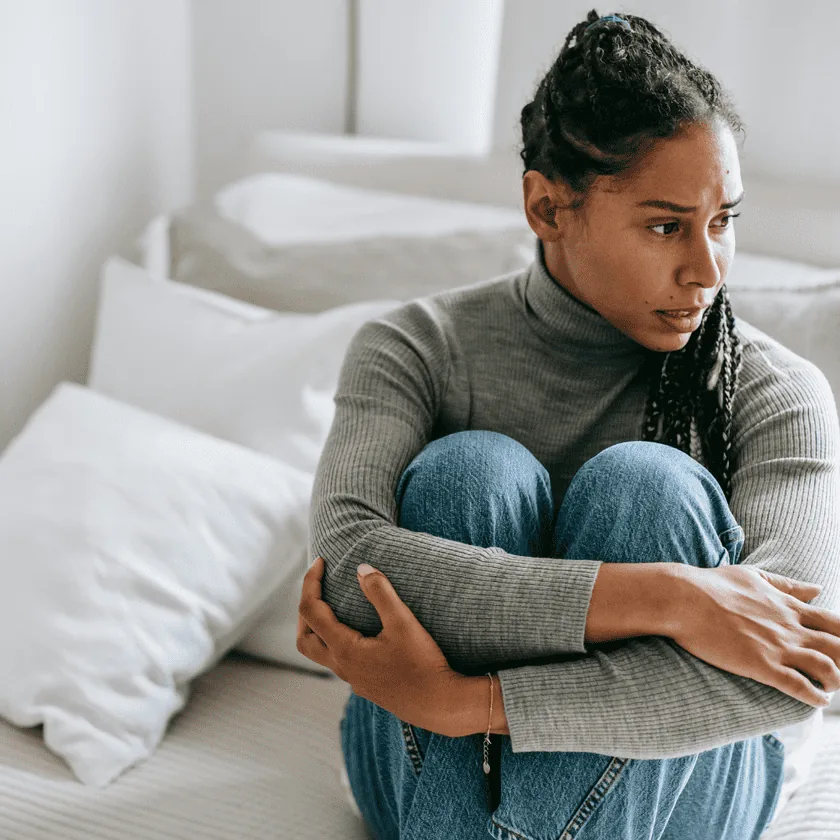 Woman sitting on a bed hugging her knees, looking worried and thoughtful