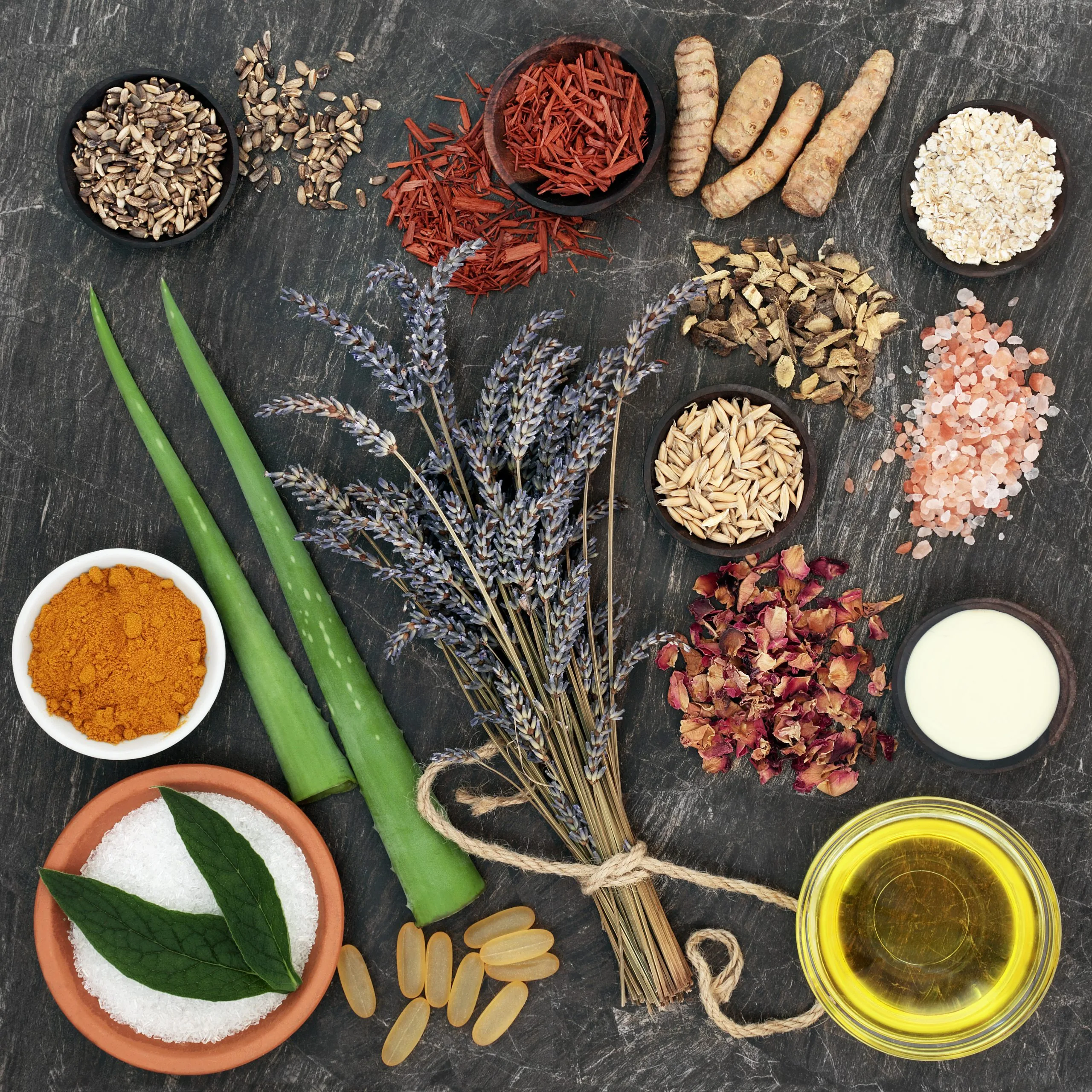 Flat lay of assorted herbs and spices—turmeric, dried lavender, aloe, oats, pink salt, oil and seeds—arranged on a dark surface.