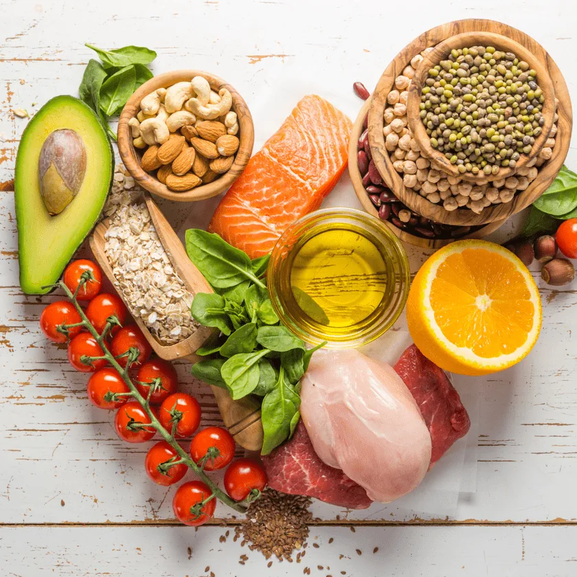 Assortment of gut-healthy foods: avocado, cherry tomatoes, nuts, oats, salmon, legumes, olive oil, orange, spinach and lean meats on a white wooden surface.