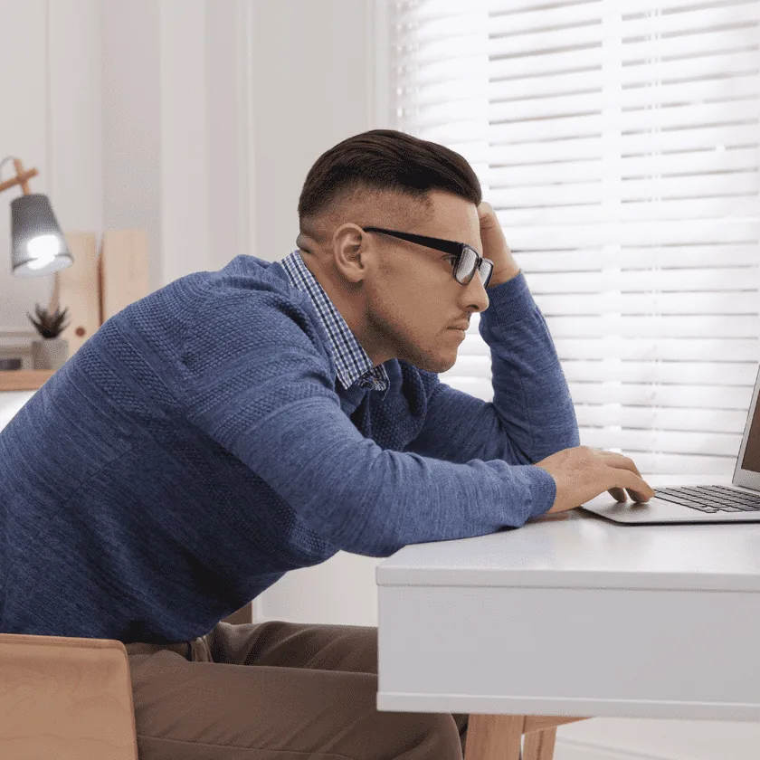 Man leaning forward with slouched posture at a desk, forehead resting on his hand while using a laptop.