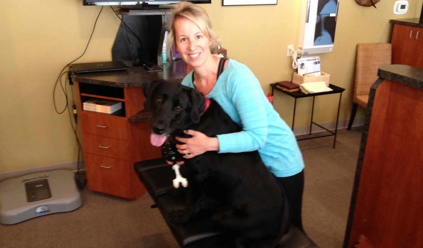 Woman hugging a black dog sitting on a clinic treatment table, the dog panting with its tongue out.