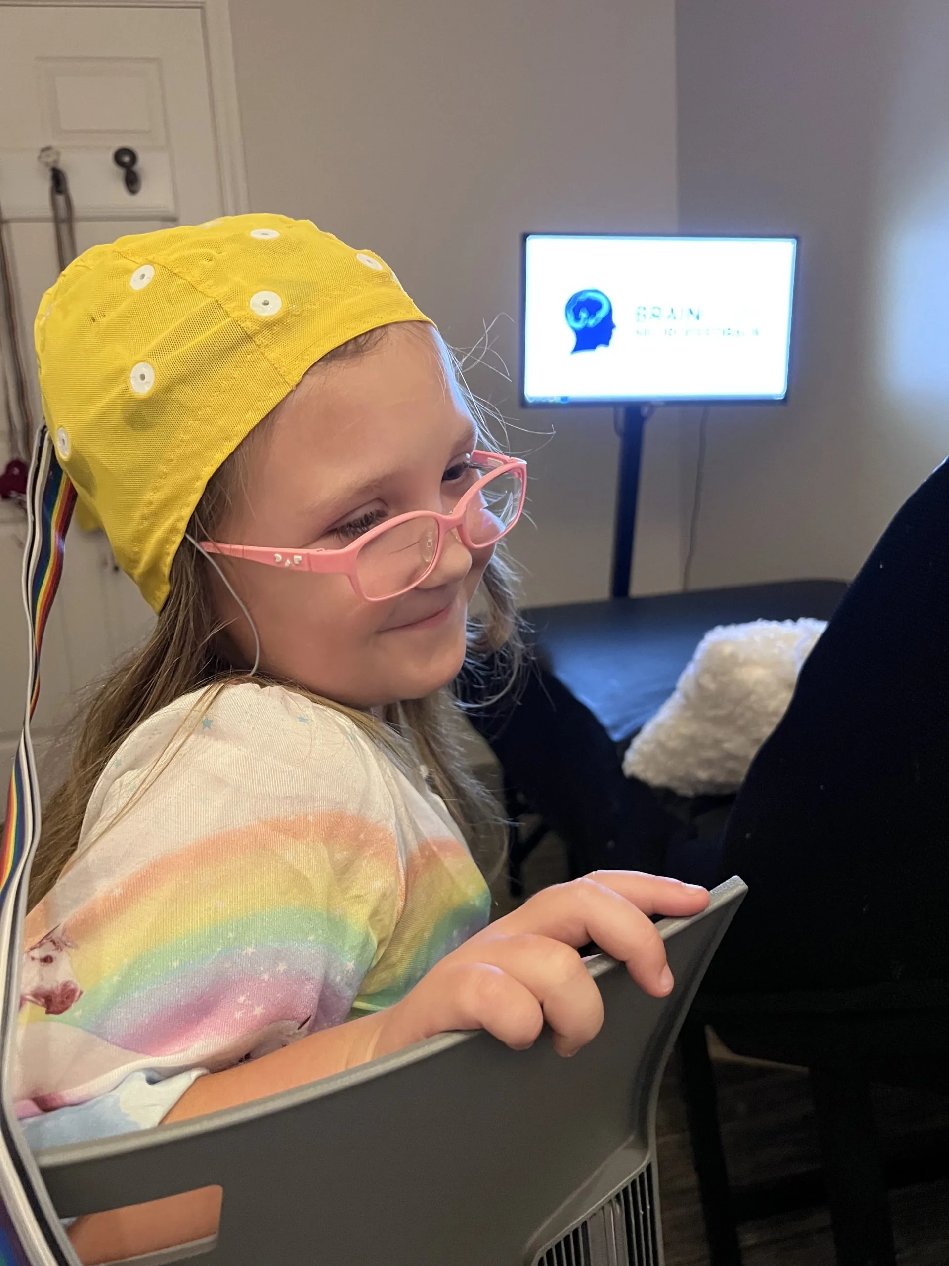 Child wearing a yellow EEG cap with sensor leads, wearing pink glasses and holding a device, with a monitor showing a brain logo in the background.