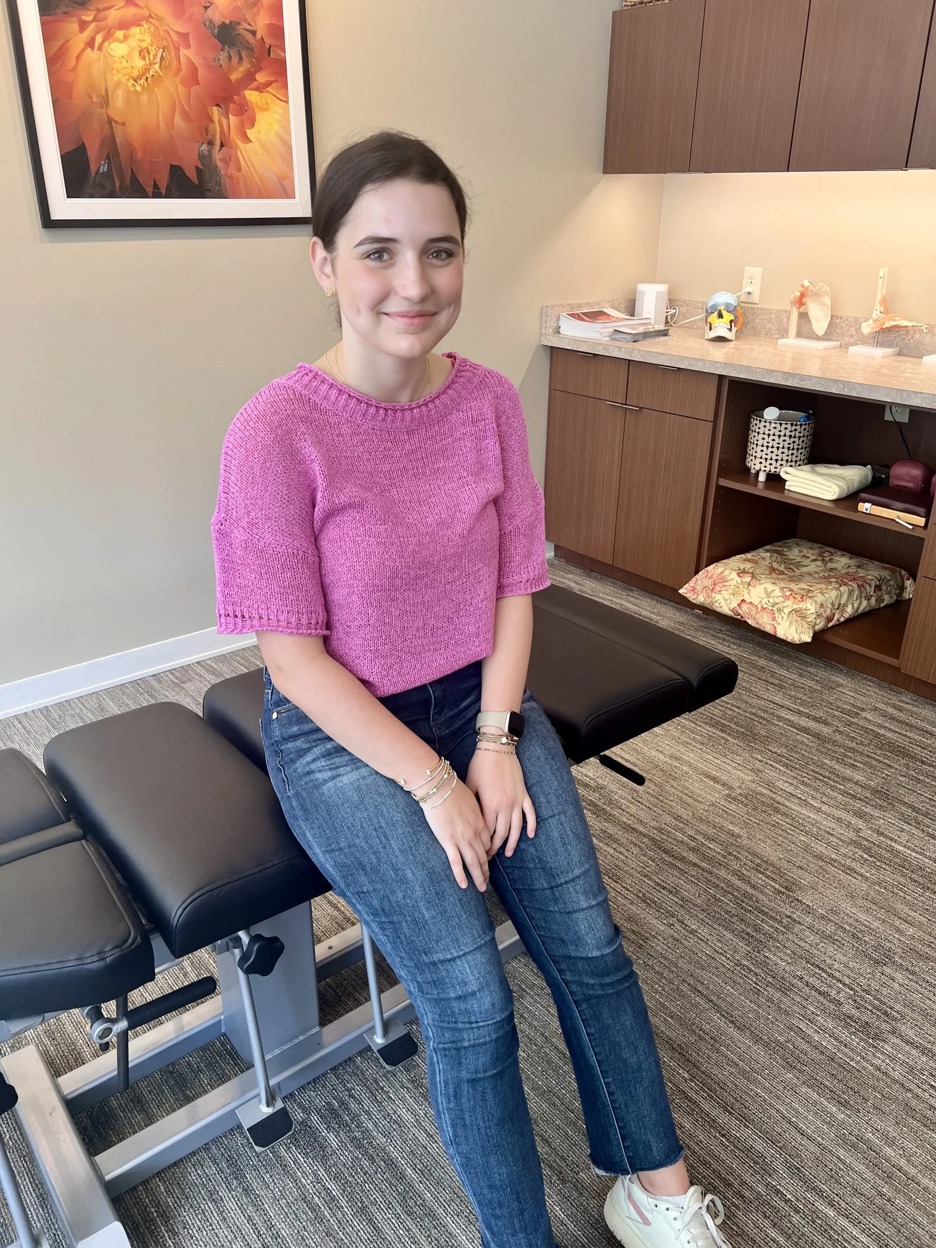 Person seated on an exam table in a clinic treatment room wearing a pink sweater; cabinets, anatomy models, and a framed floral print visible.