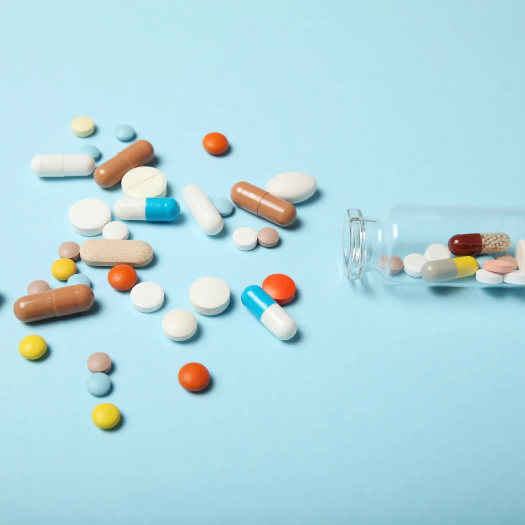 Various prescription and over-the-counter pills and capsules spilling from a small glass bottle onto a light blue surface