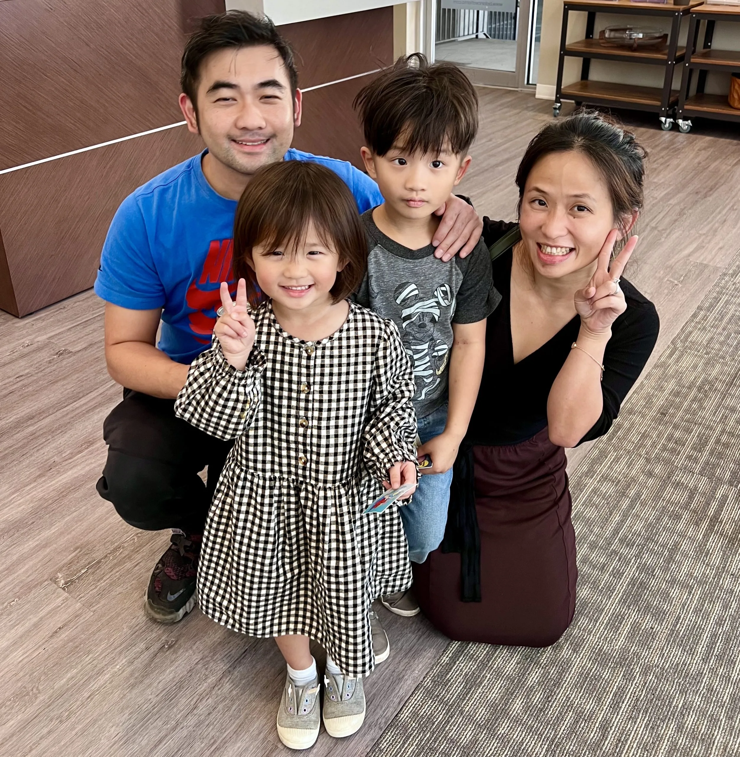 Two adults kneel behind two children holding up peace signs in a clinic reception area.