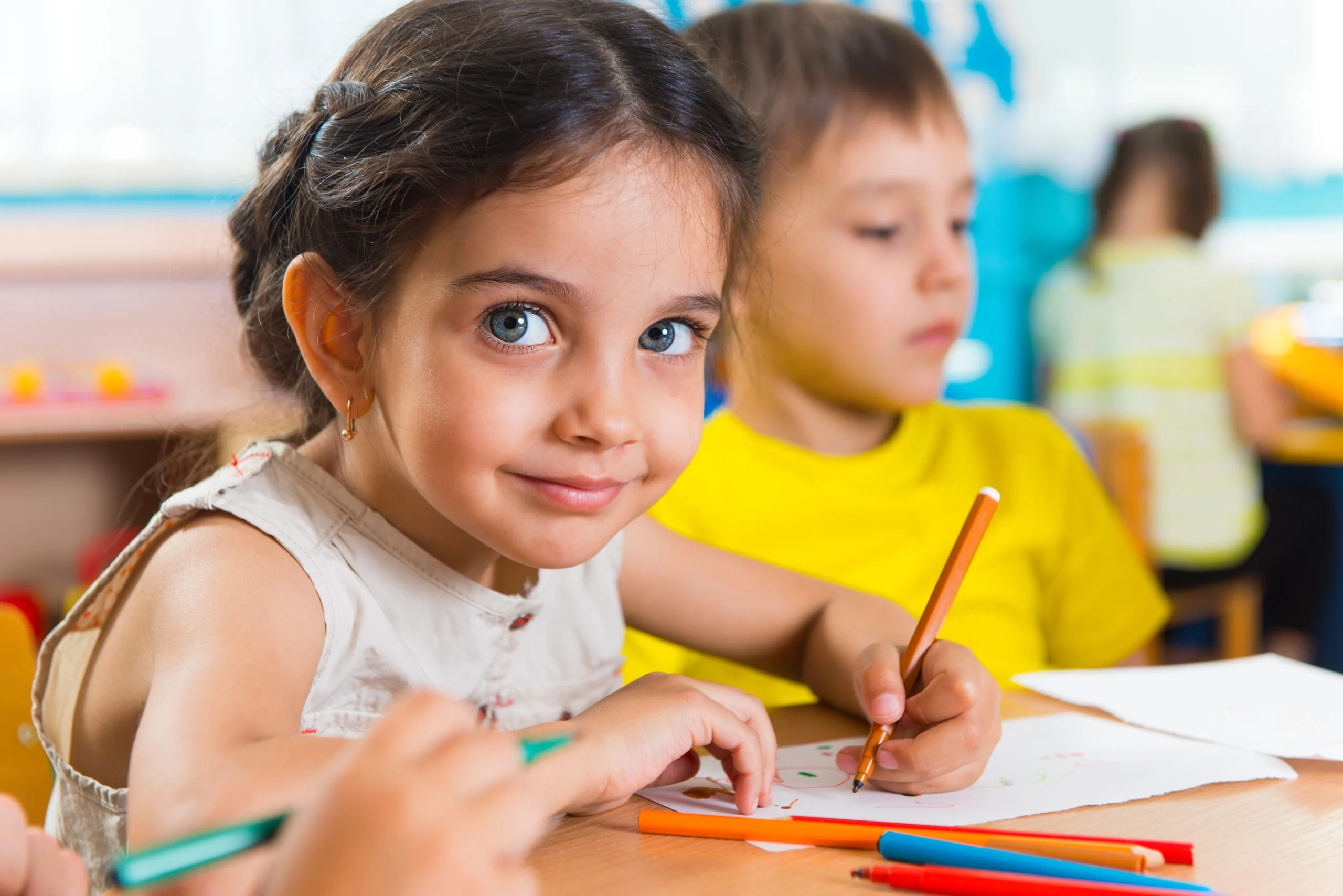 Girl with wide eyes drawing with a pencil at a classroom table, colored markers on the table and another child blurred in the background.