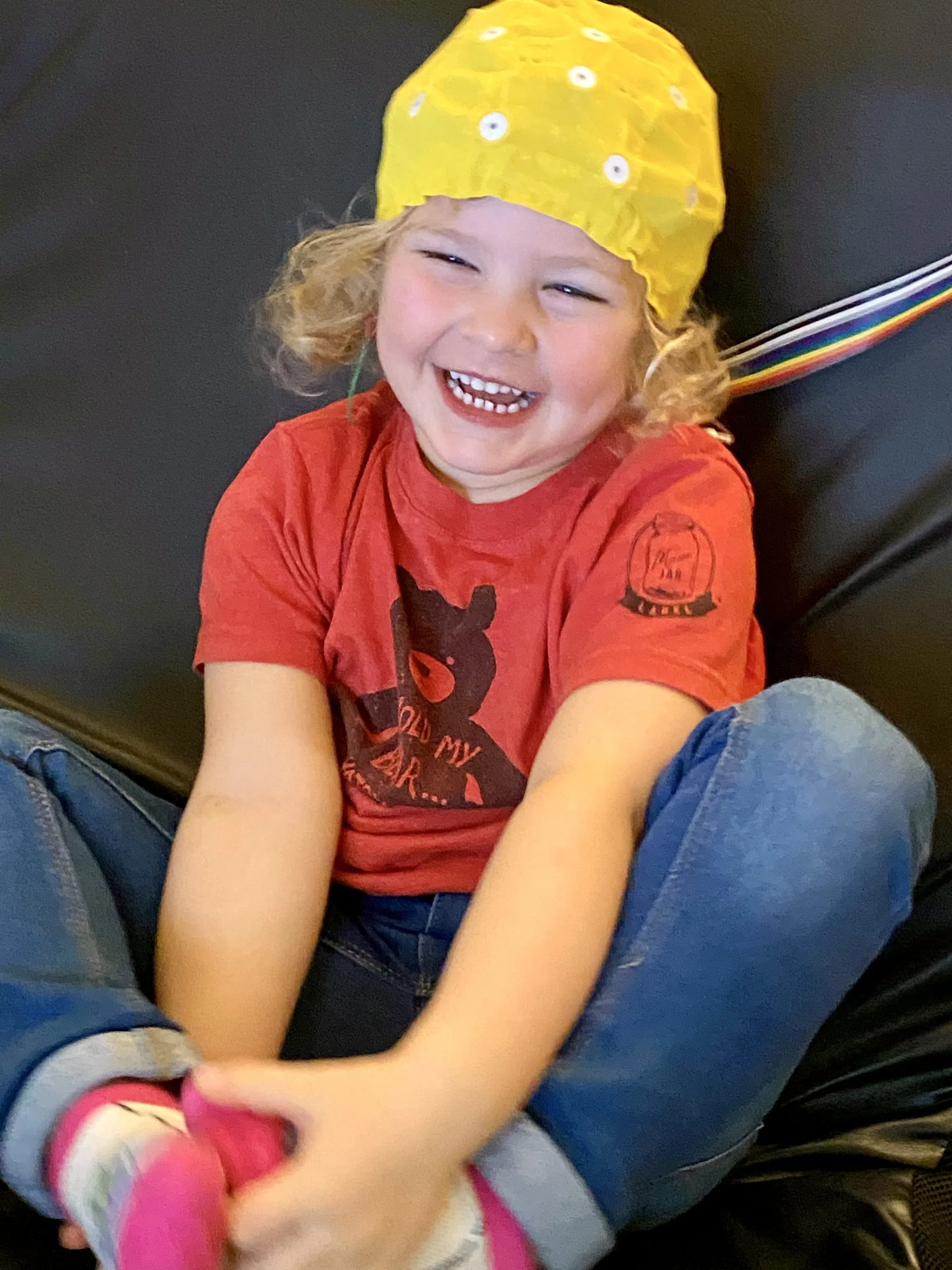 Child wearing a yellow EEG cap and smiling while seated during a brain activity assessment