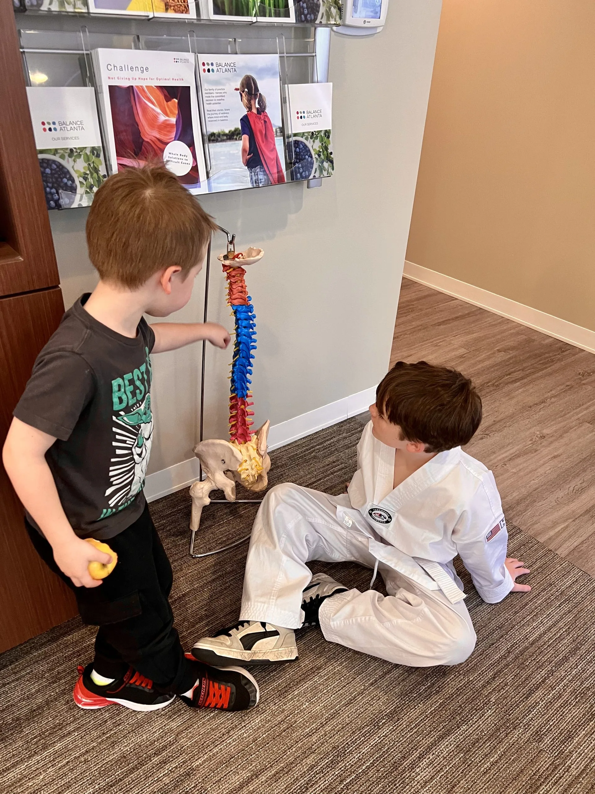 Two boys examine a colorful anatomical spine model in a clinic waiting area with brochures on the wall