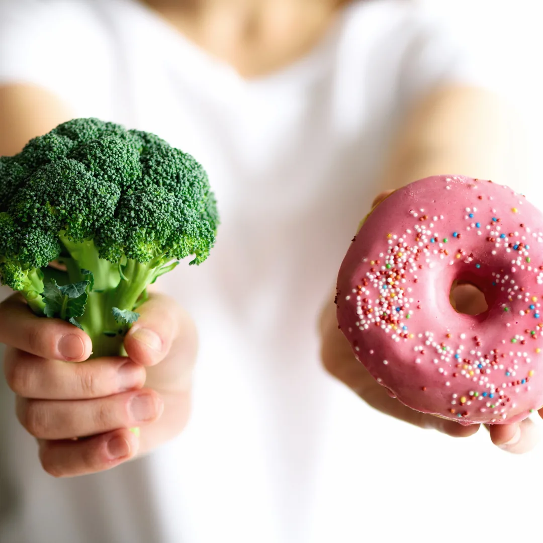 Hands holding a broccoli floret in one hand and a pink-frosted donut with sprinkles in the other against a blurred white background.
