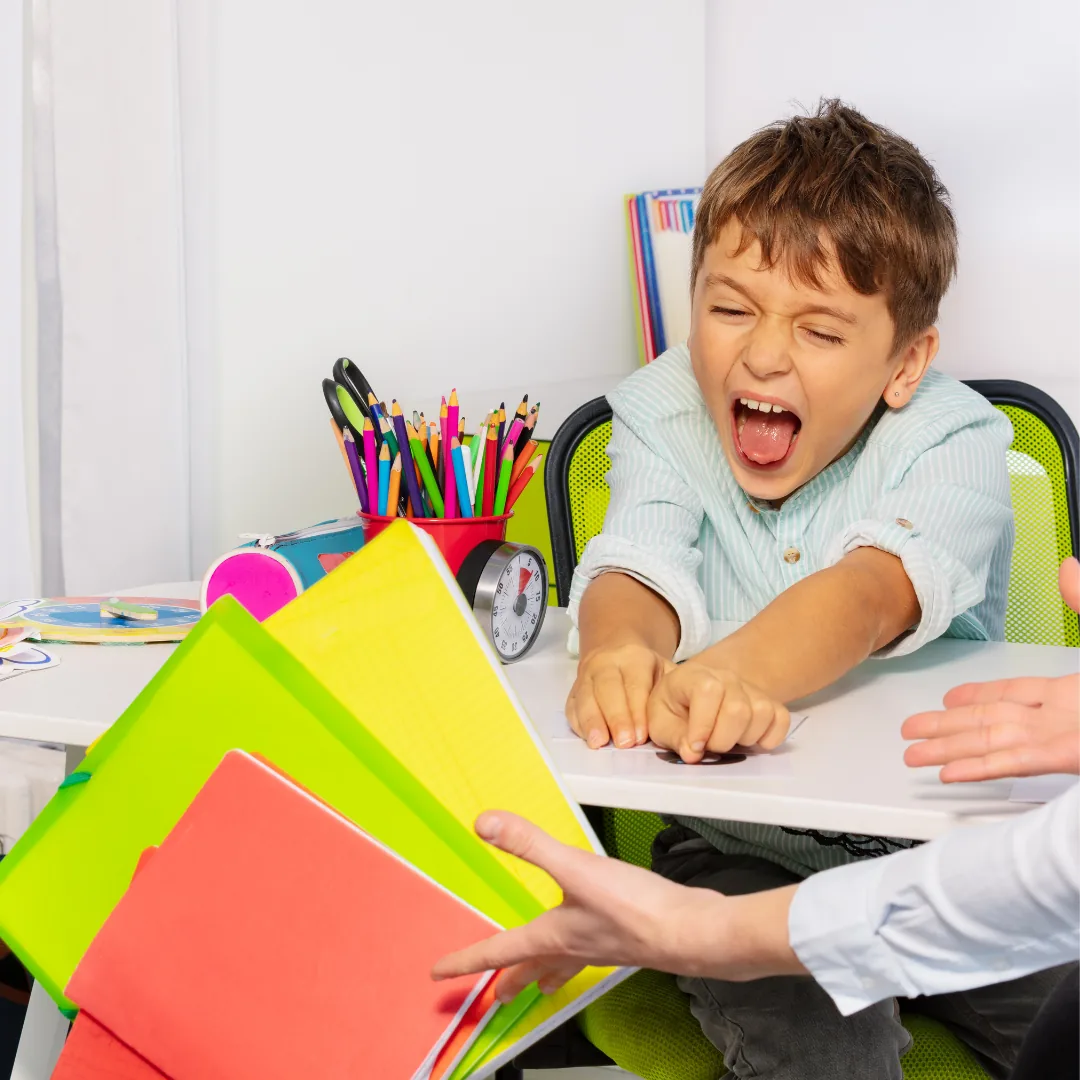 Child at a desk shouting with mouth wide open while reaching toward colorful notebooks and school supplies.