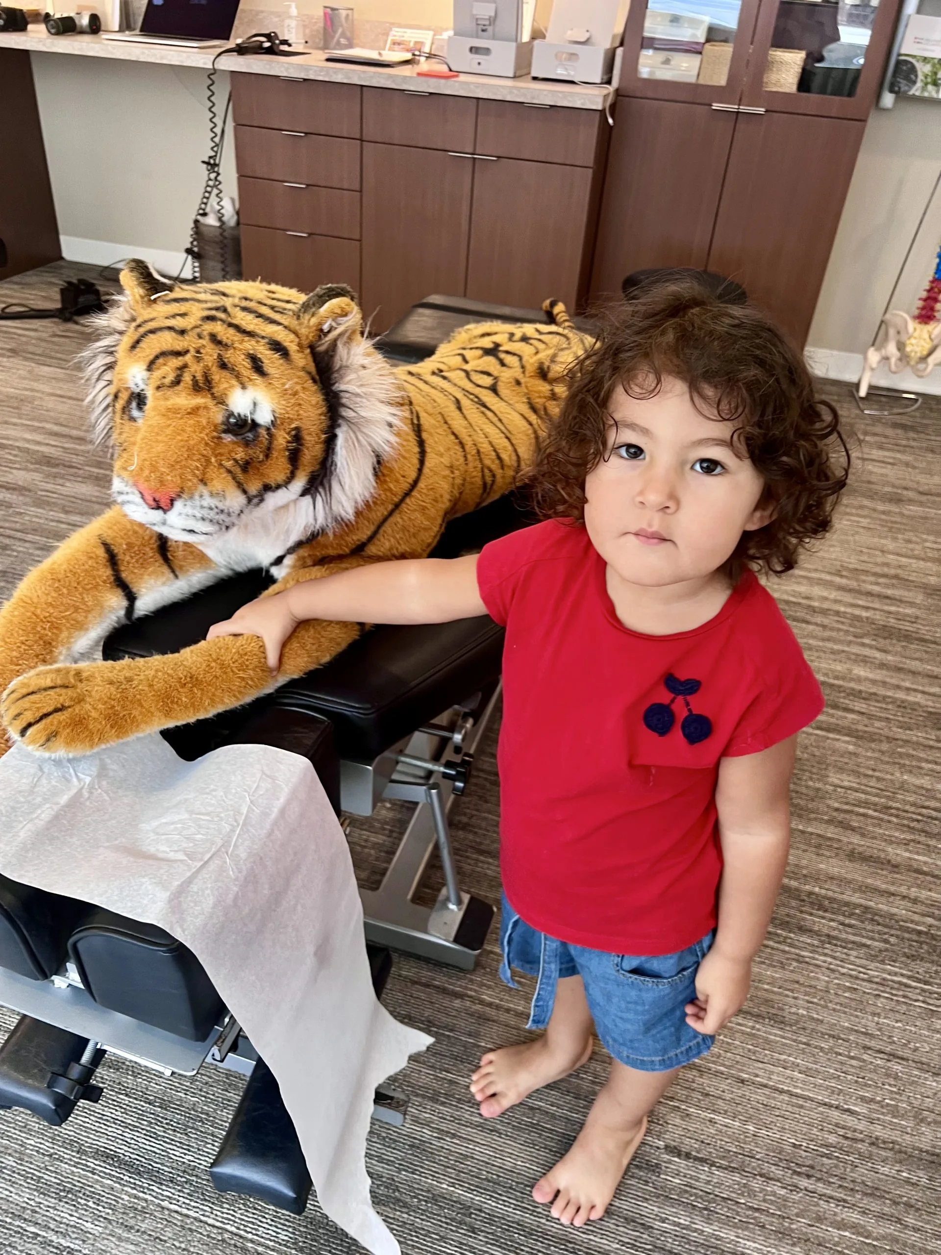 Child in a red shirt standing barefoot beside a large stuffed tiger on a clinic exam table.