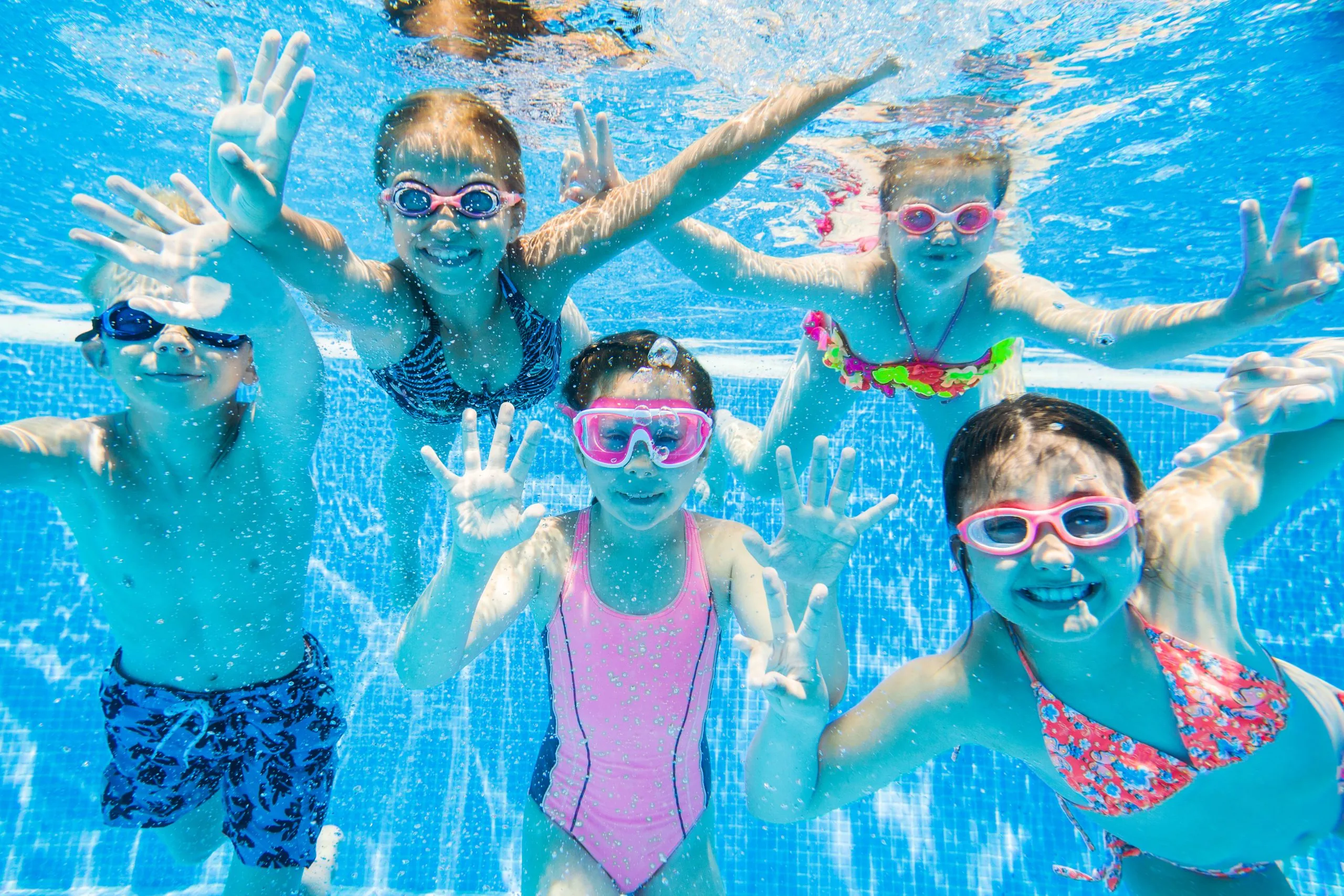 children wearing goggles swimming underwater in a pool, reaching toward the camera