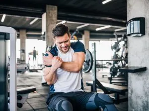 Man seated on a gym bench clutching his upper arm near the shoulder after a workout