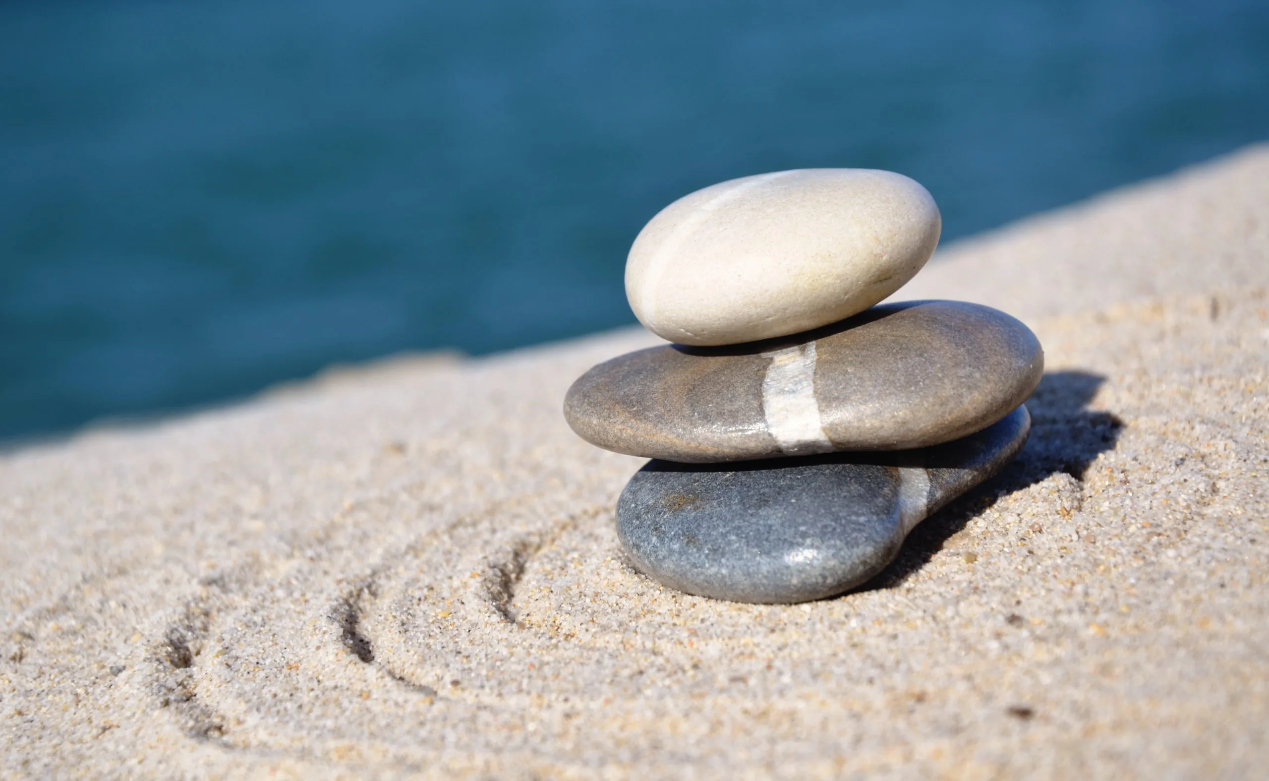 Three smooth stones stacked on sand with blurred blue water behind, a calm composition suggesting balance and recovery for concussion/head trauma