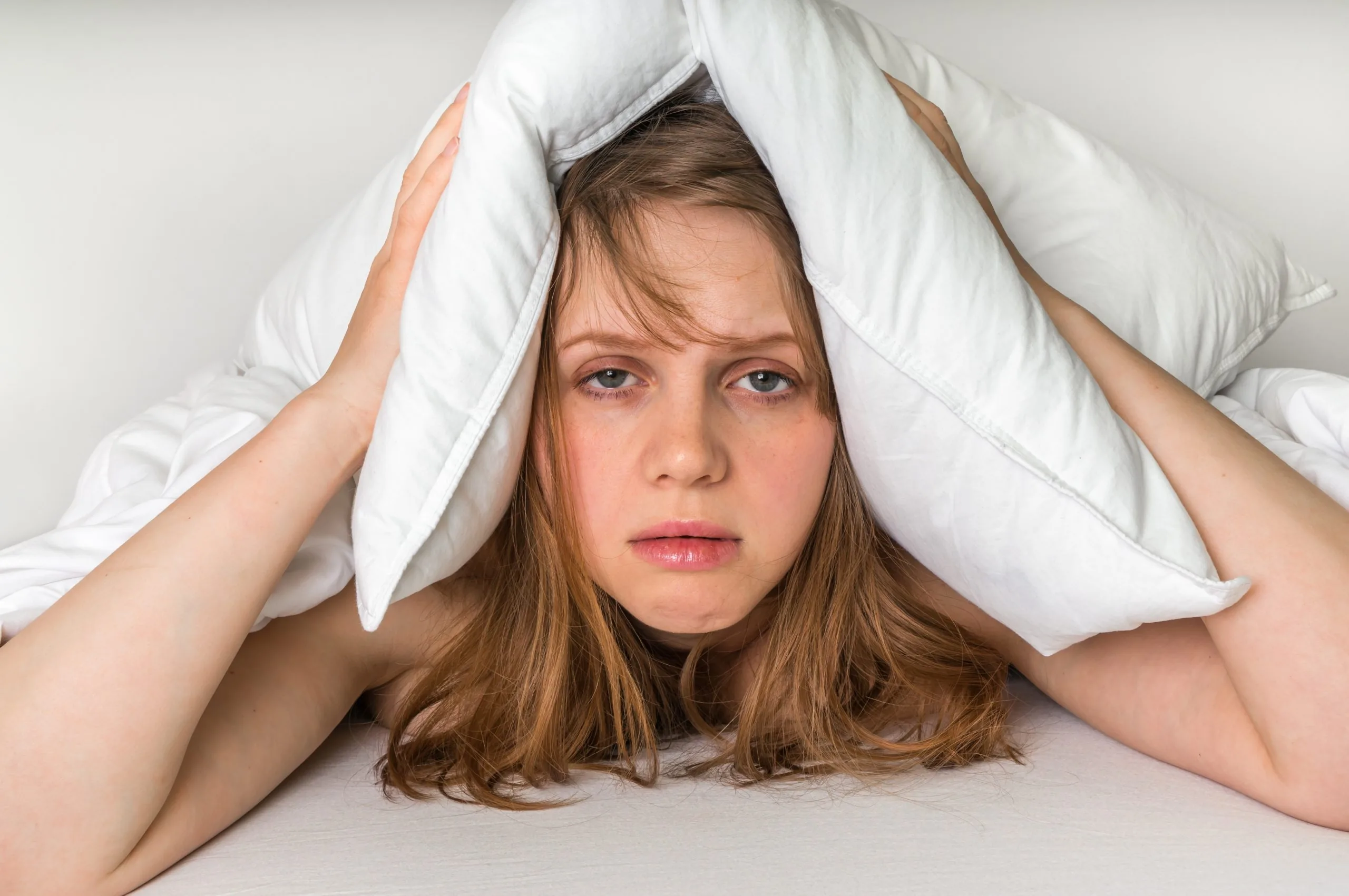 Woman lying down holding a white pillow over her head with both hands, eyes partially open.