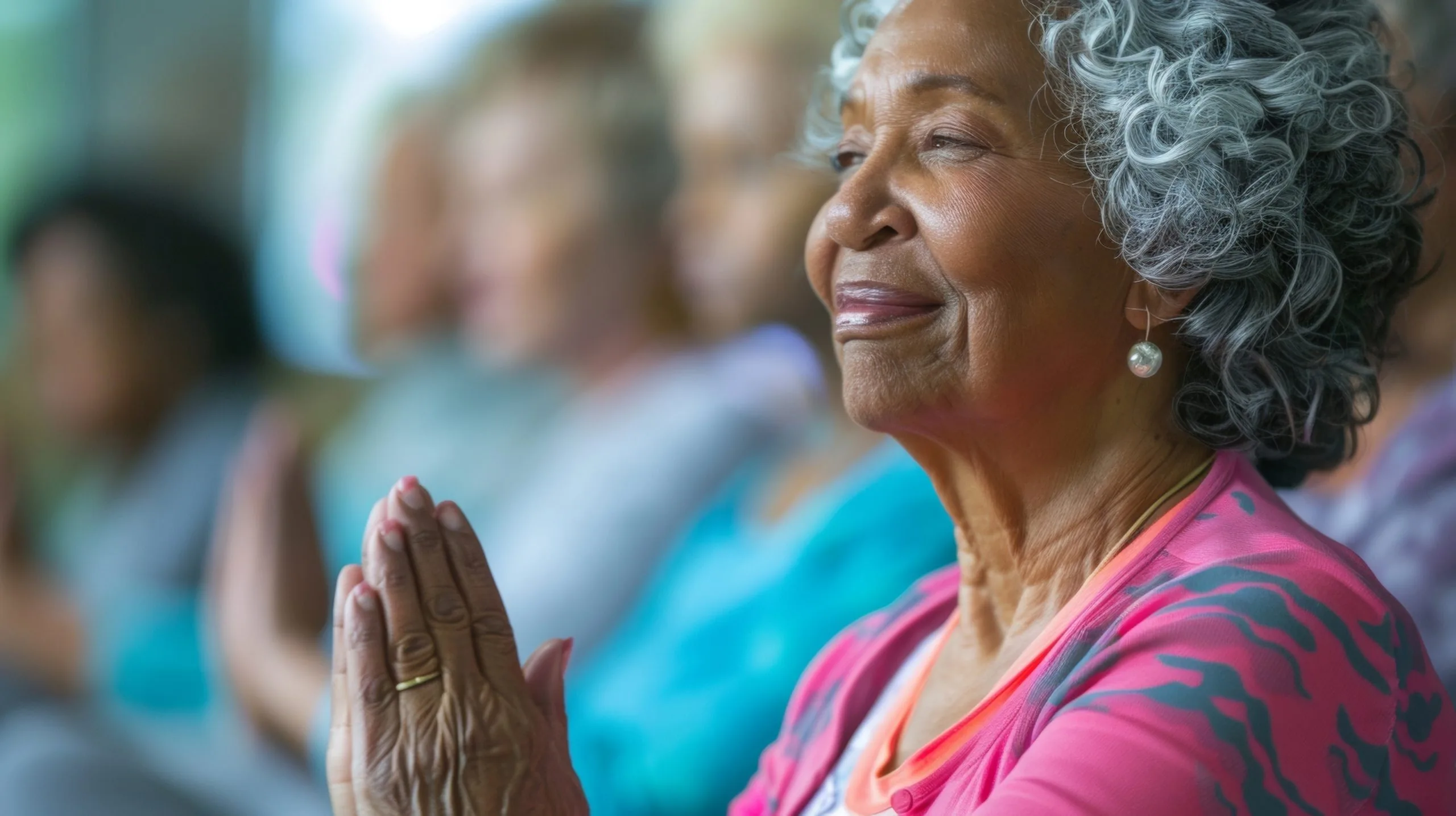 Older woman smiling with hands pressed together in a gentle meditative pose, other participants blurred in the background