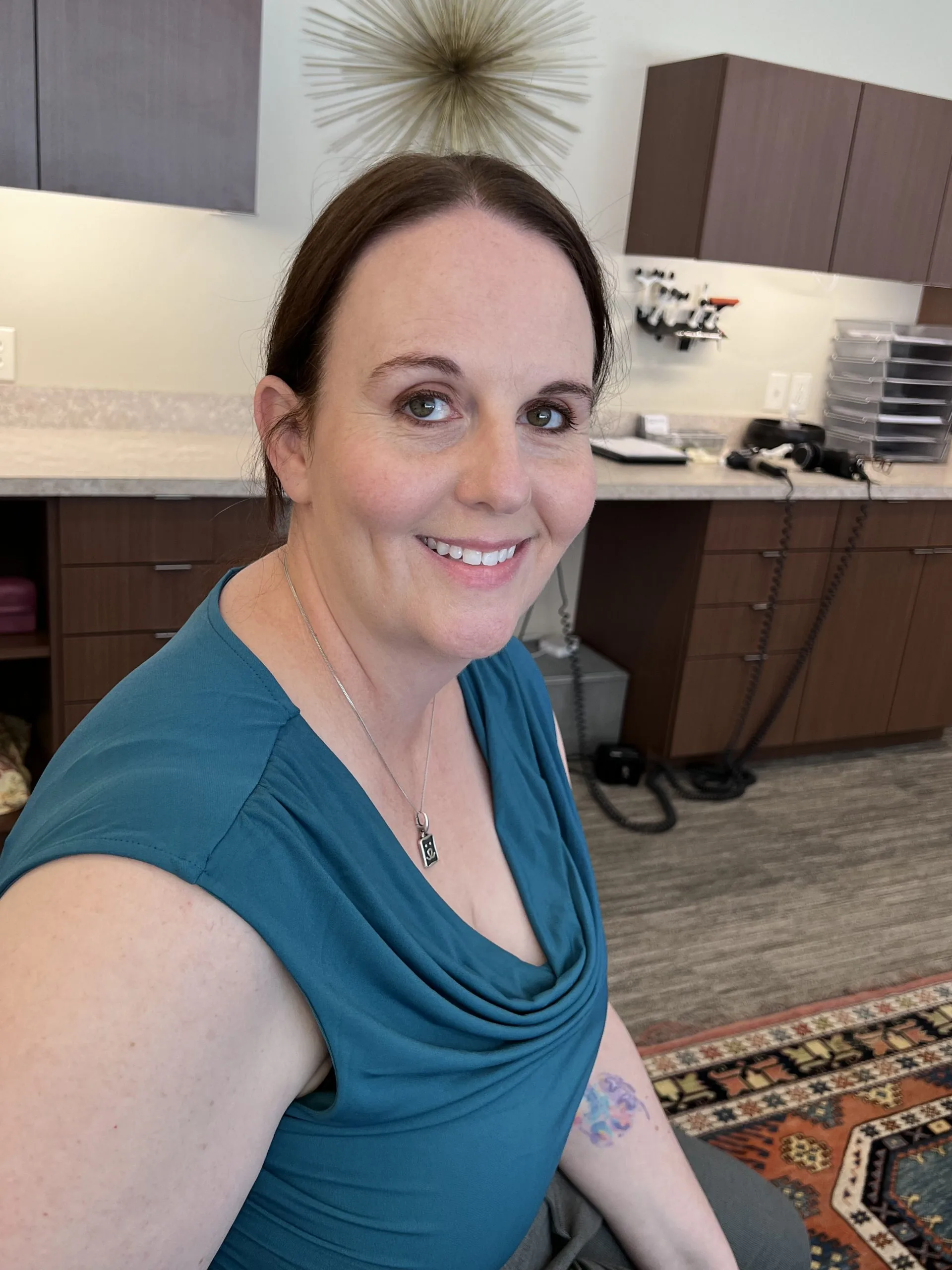 Woman seated in a chiropractic exam room showing her lower face and jaw, smiling with cabinets and clinical equipment visible behind her.