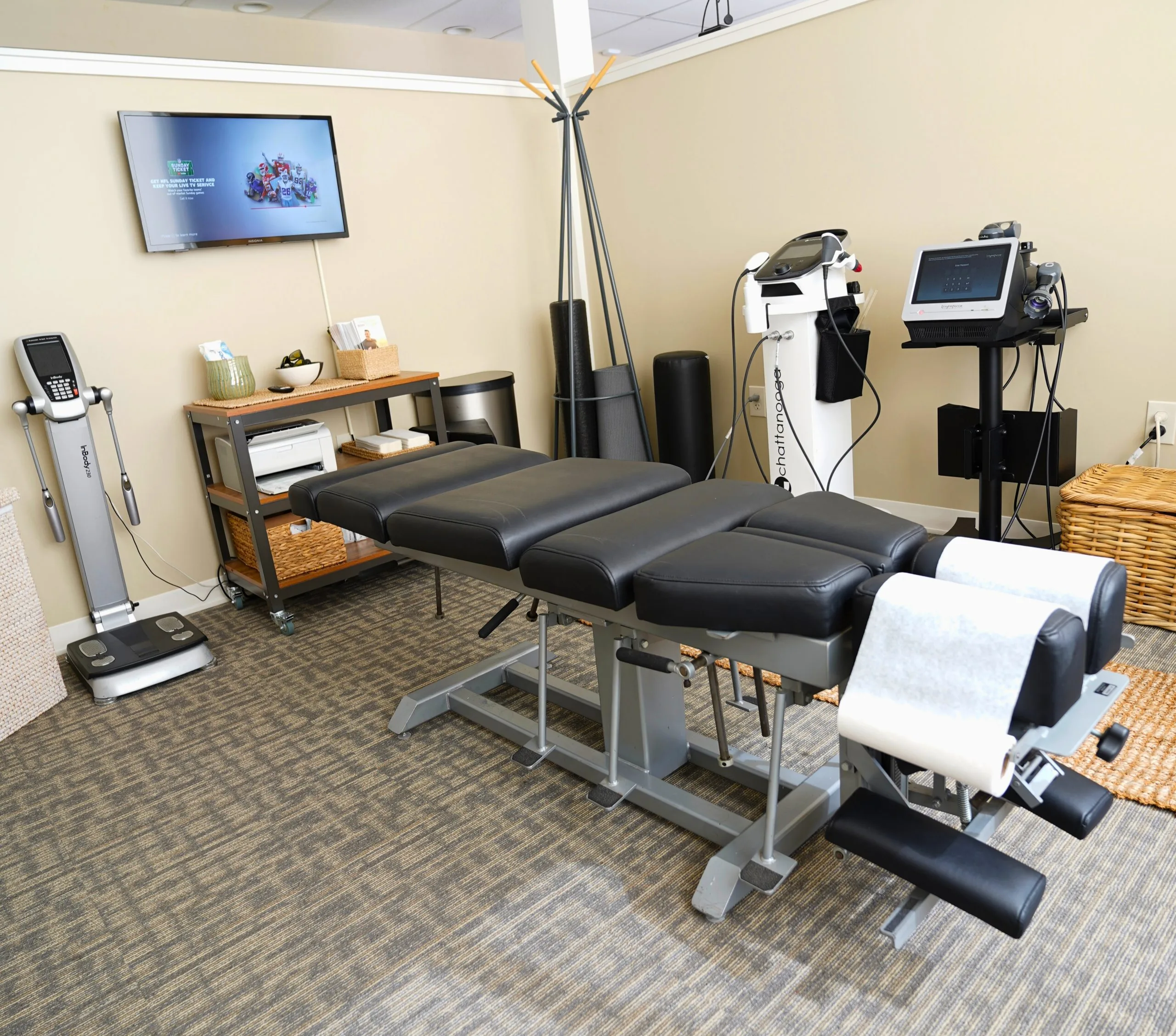 Chiropractic treatment table with therapeutic machines, foam rollers and storage shelves in a clinic room for muscle-spasm care.