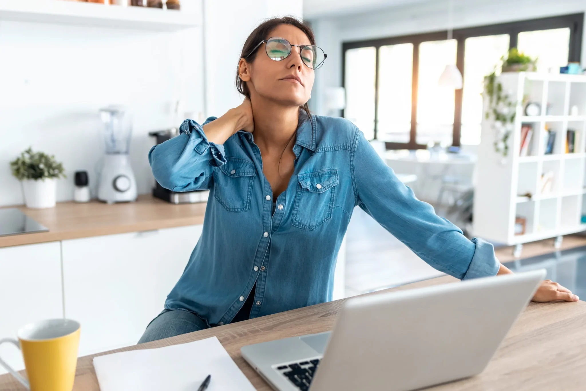 Woman at a kitchen counter holding the back of her neck with one hand, indicating neck pain or stiffness.