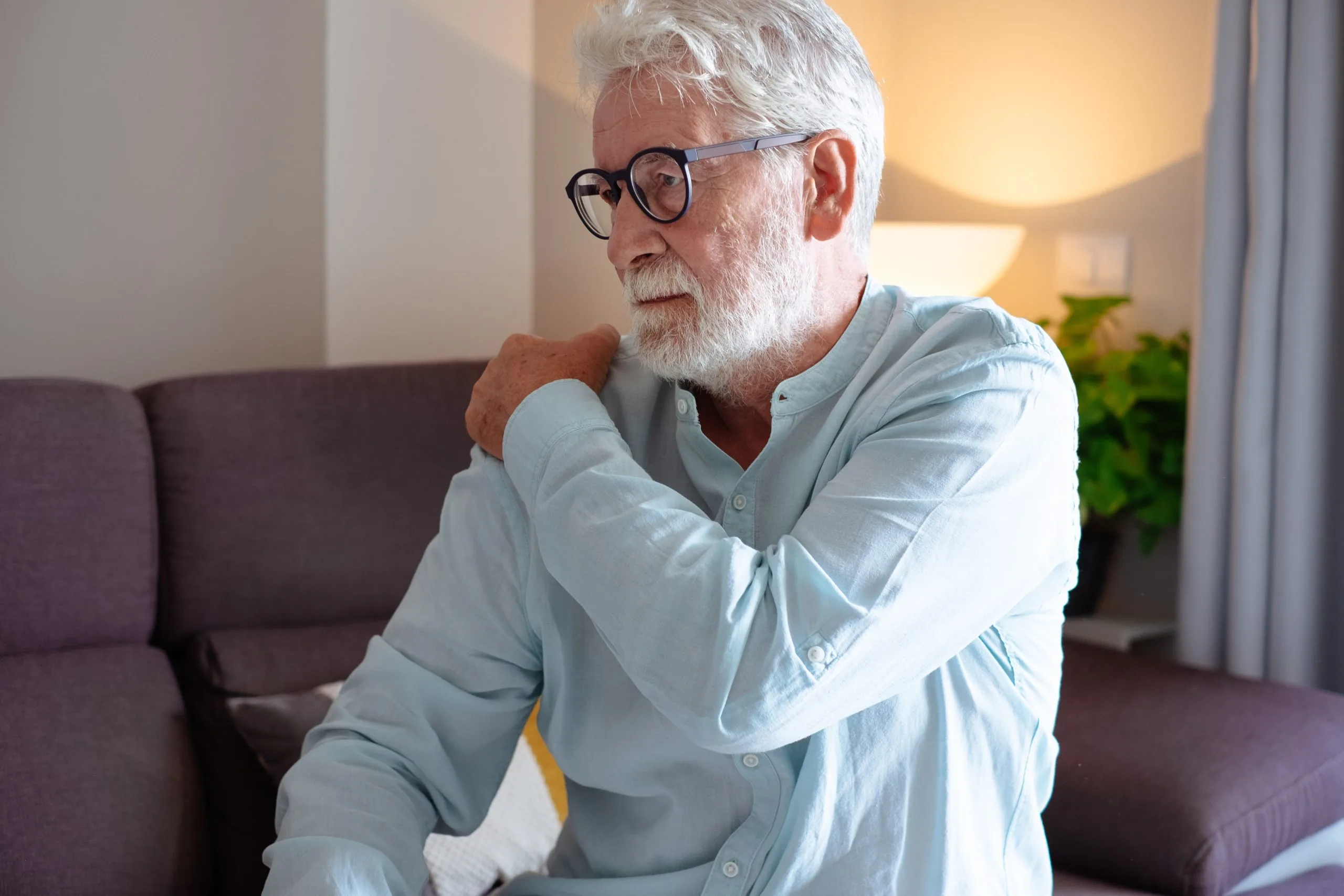 Man seated on a couch holding his right shoulder and neck while wearing glasses and a light blue shirt