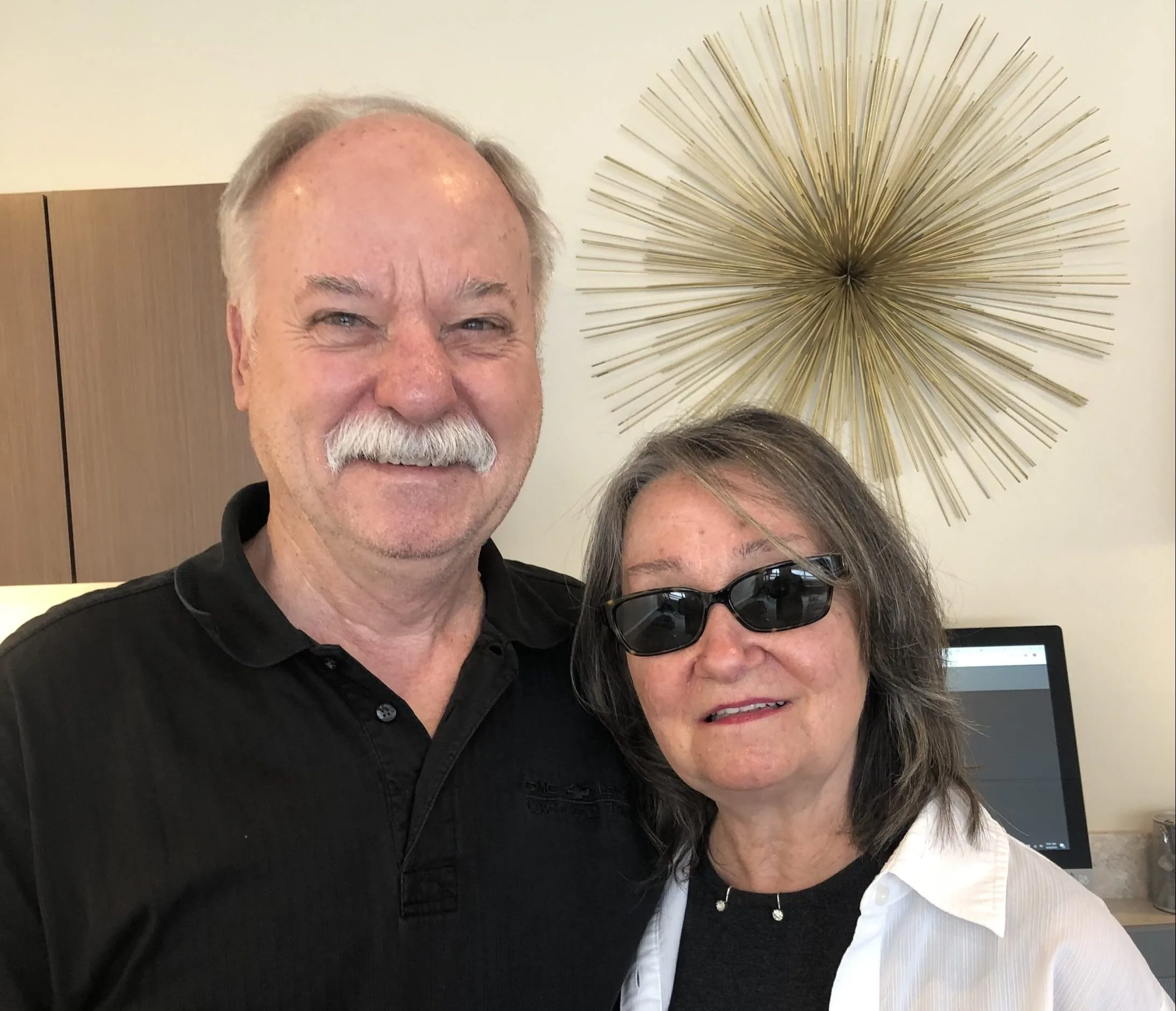 Two people taking a close selfie in a clinic reception area with decorative starburst wall art during a chiropractic visit.