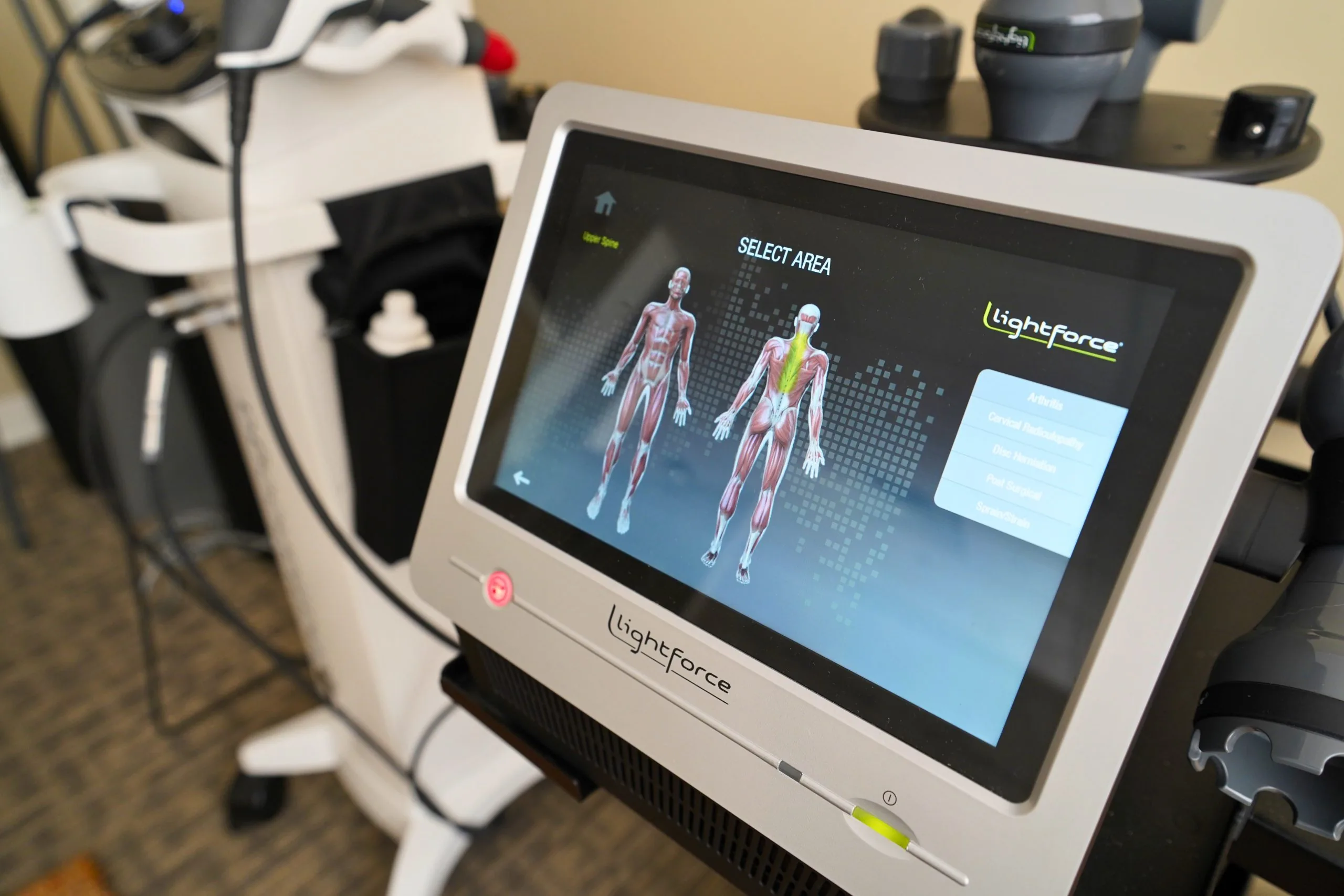 Laser therapy console showing anatomical muscle diagrams and 'Select Area' menu, with the handheld laser wand visible for shoulder-pain treatment.