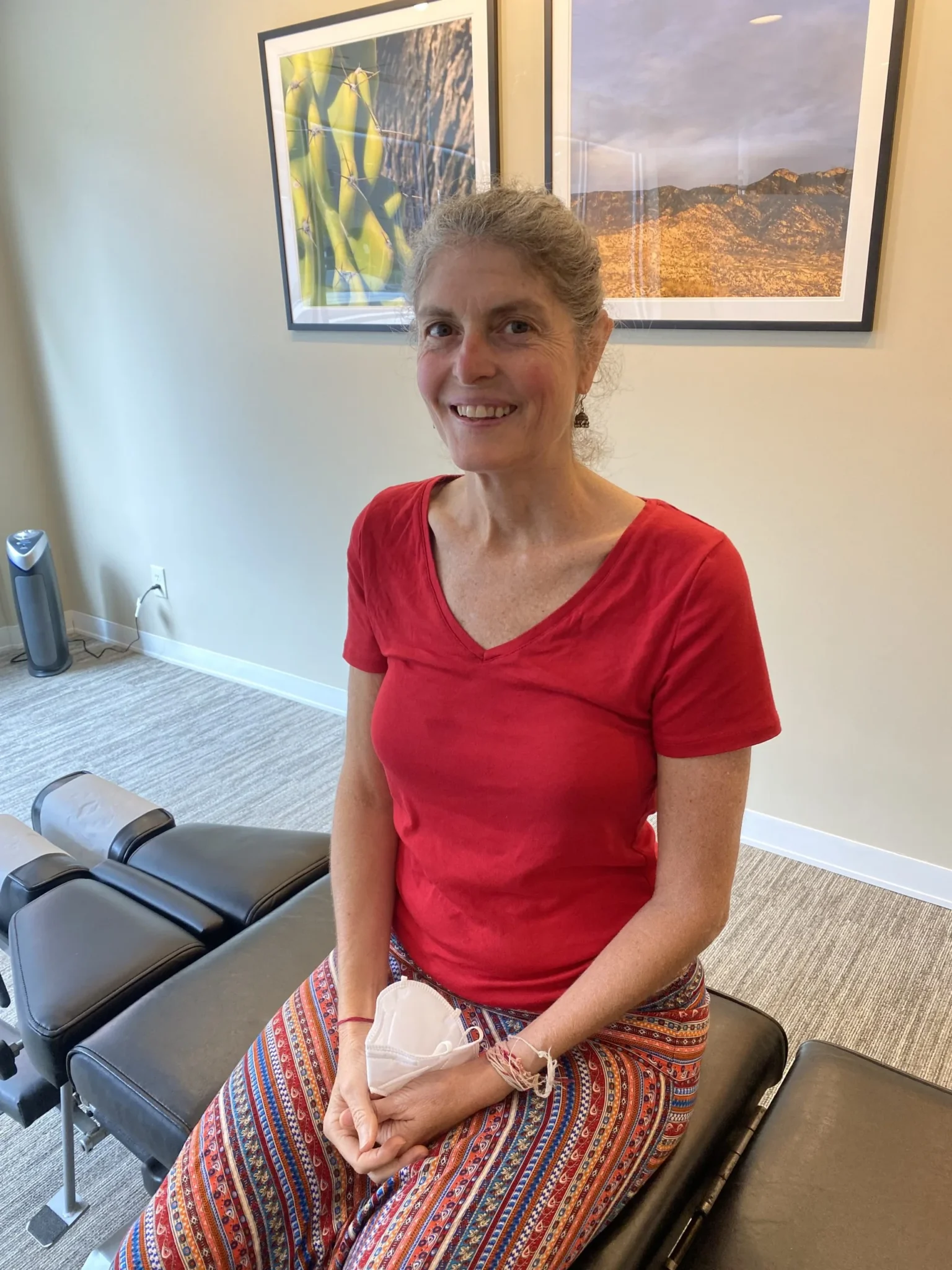 Person seated on a chiropractic treatment table holding a white face mask in a clinic room with framed artwork behind