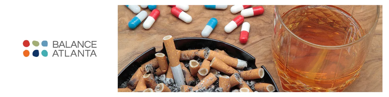 Ashtray full of cigarette butts, scattered red-and-white capsules, and a glass of alcohol on a wooden surface beside the Balance Atlanta logo.