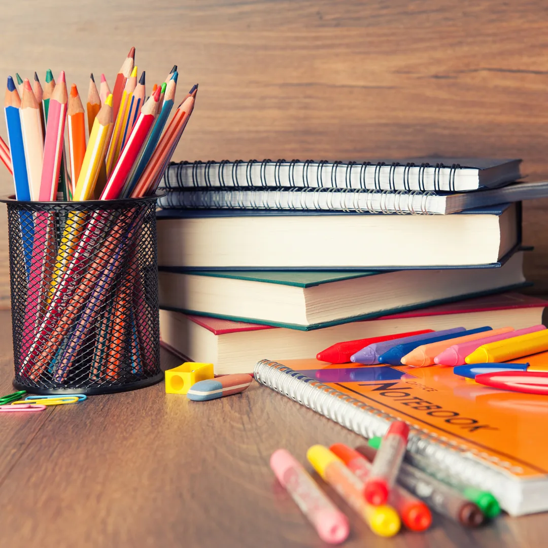 Metal pencil cup filled with colored pencils beside stacked spiral notebooks and scattered markers and crayons on a wooden desk.