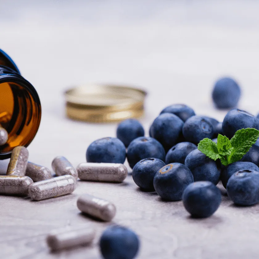 Blueberries and herbal supplement capsules spilled from an amber bottle, with a small mint sprig on a light tabletop.