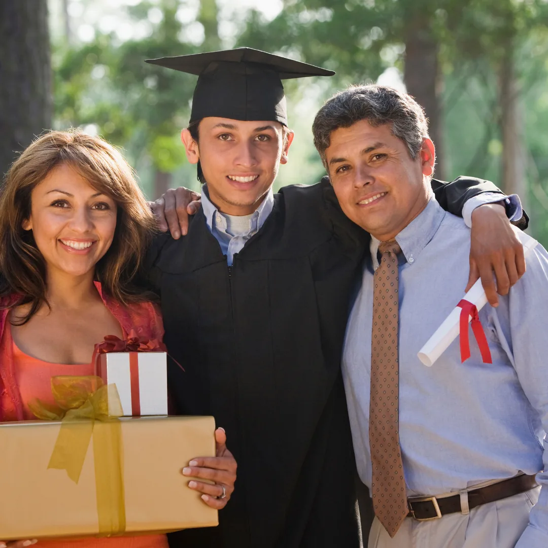 Graduate in cap and gown stands between smiling parents; one parent holds wrapped gifts and the other holds a rolled diploma tied with a red ribbon.