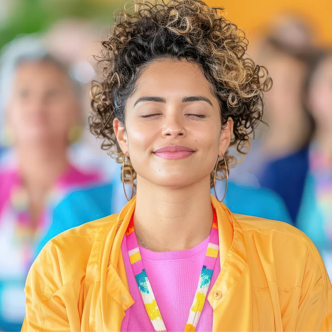 Person with eyes closed wearing a yellow jacket, pink top, and a colorful beaded lanyard in a blurred group setting