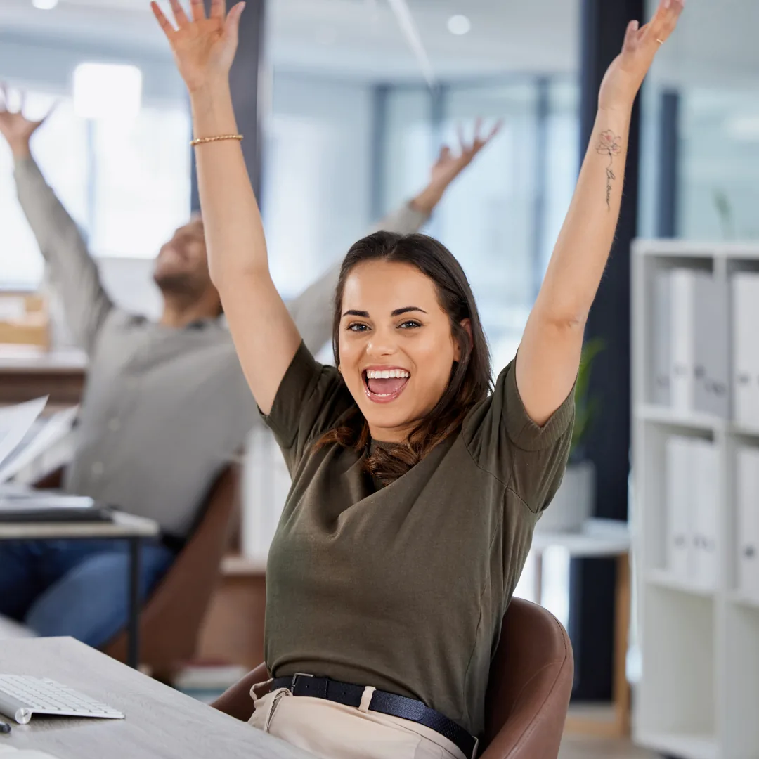 Smiling woman seated at an office desk with both arms raised overhead.