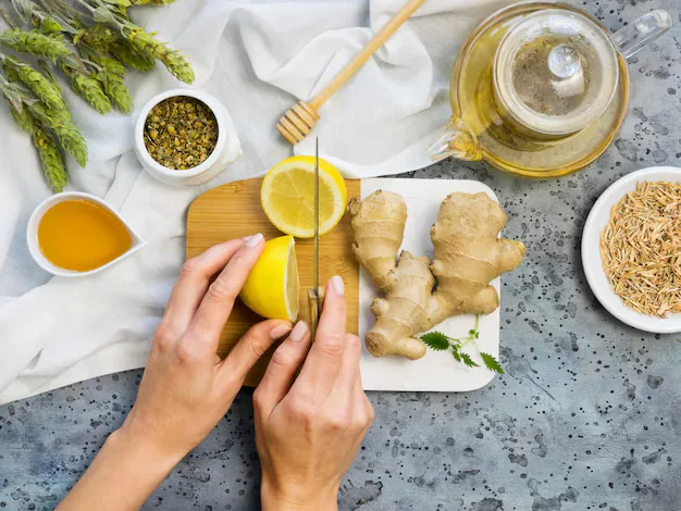 Hands slicing a lemon on a cutting board beside fresh ginger, honey, chamomile, and a teapot, arranged as natural remedies for allergies.