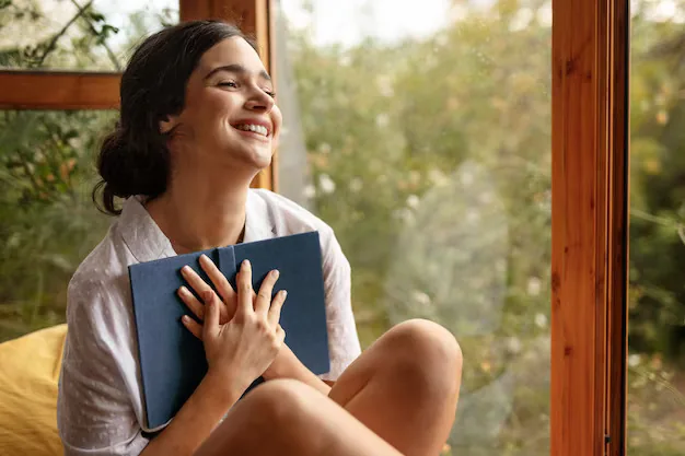 Smiling person holding a closed blue book to their chest while sitting by a window.