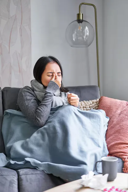 Woman wrapped in a blanket on a couch holding a tissue to her nose, appearing to sneeze and have nasal congestion.