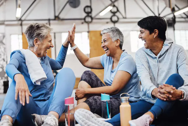 Three women on a gym floor laughing and high-fiving with water bottles nearby, suggesting supportive group wellness for allergy care.
