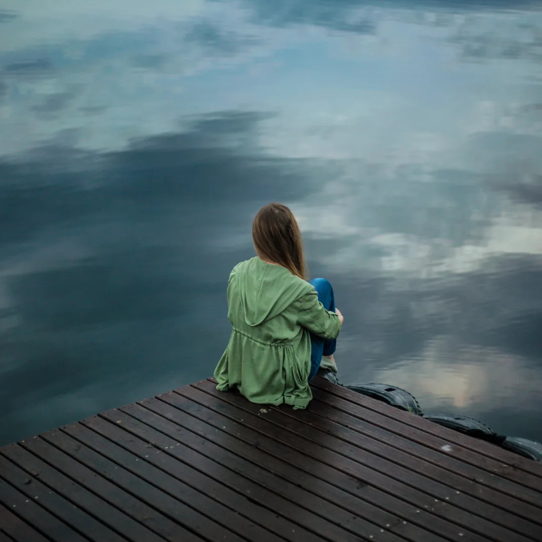 Person sitting on a wooden dock with knees drawn to chest, seen from behind, looking out over calm reflective water.