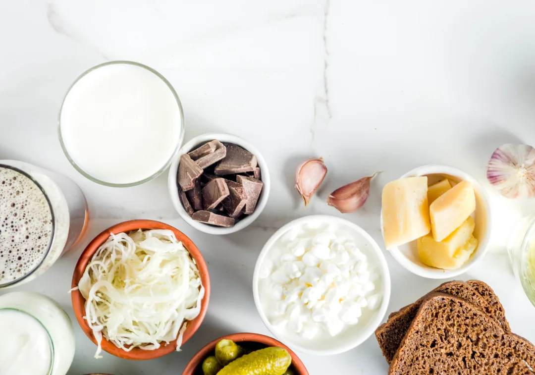 Assorted probiotic and fermented foods on a white surface, including sauerkraut, cottage cheese, pickles, milk, cheese chunks, dark bread, chocolate, and garlic.