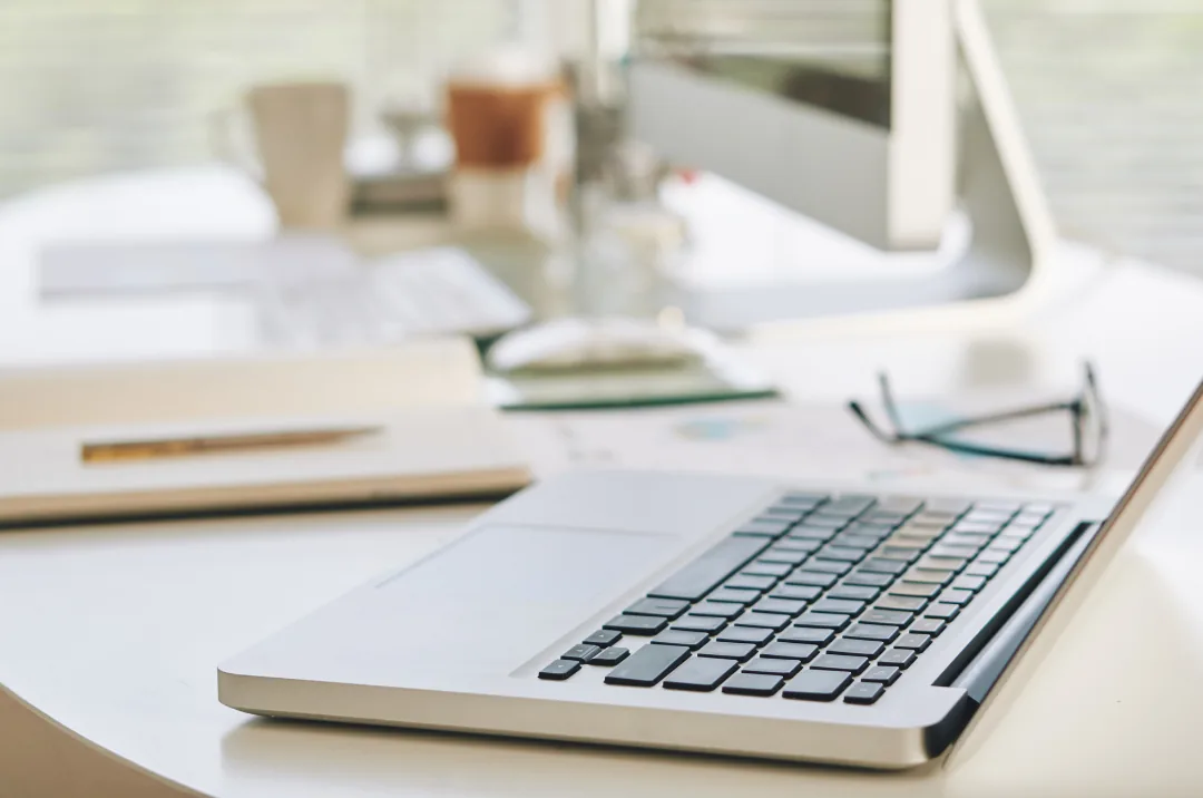 Laptop with glasses and notebooks on a bright desk, monitor blurred in the background.
