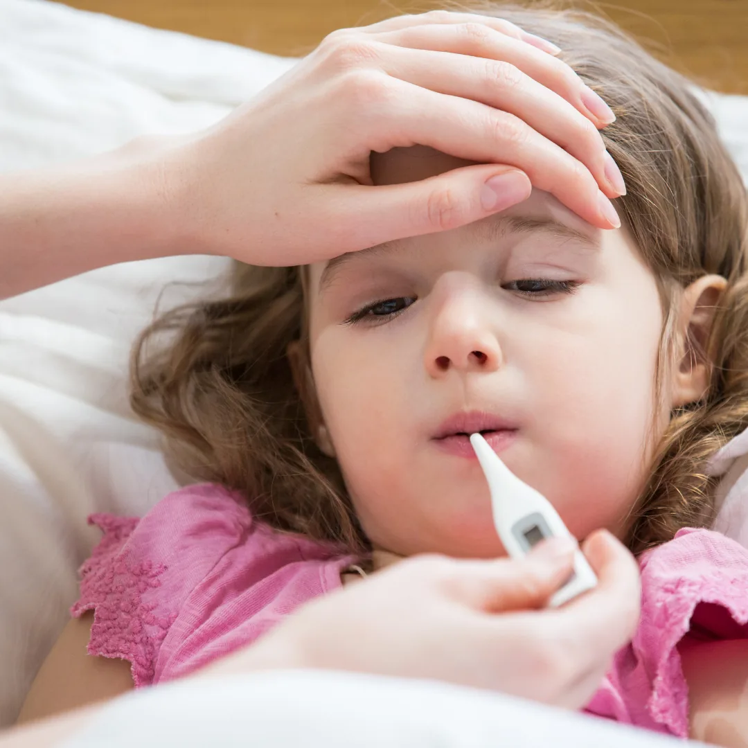 Child lying in bed having a digital thermometer placed in their mouth while a caregiver holds their forehead.