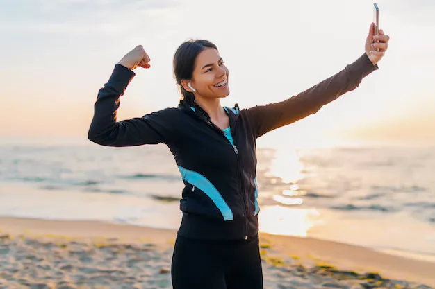 Woman smiling and flexing her arm while taking a selfie on a beach at sunrise, wearing workout clothes and earbuds.
